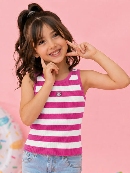 A young girl with dark brown hair in a high ponytail poses in a pink and white striped ribbed tank top and light blue jeans against a solid pink background.