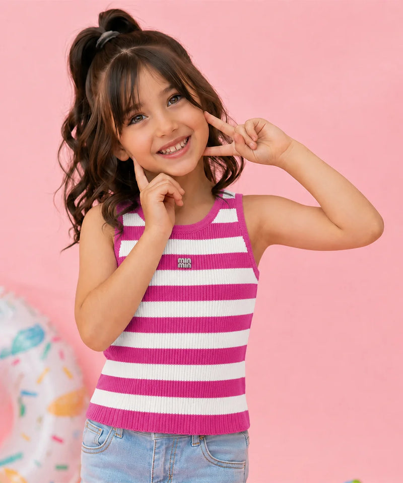 A young girl with dark brown hair in a high ponytail poses in a pink and white striped ribbed tank top and light blue jeans against a solid pink background.
