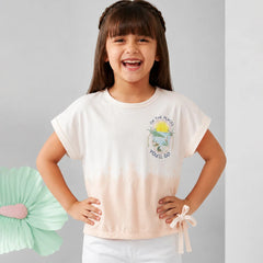 A young girl with dark hair in braids smiling, wearing a short-sleeve tie-dye t-shirt with a small graphic on the chest and white pants, standing against a light gray background.