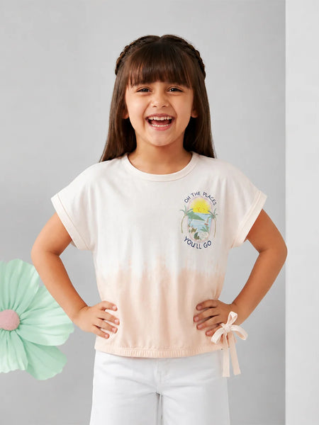 A young girl with dark hair in braids smiling, wearing a short-sleeve tie-dye t-shirt with a small graphic on the chest and white pants, standing against a light gray background.