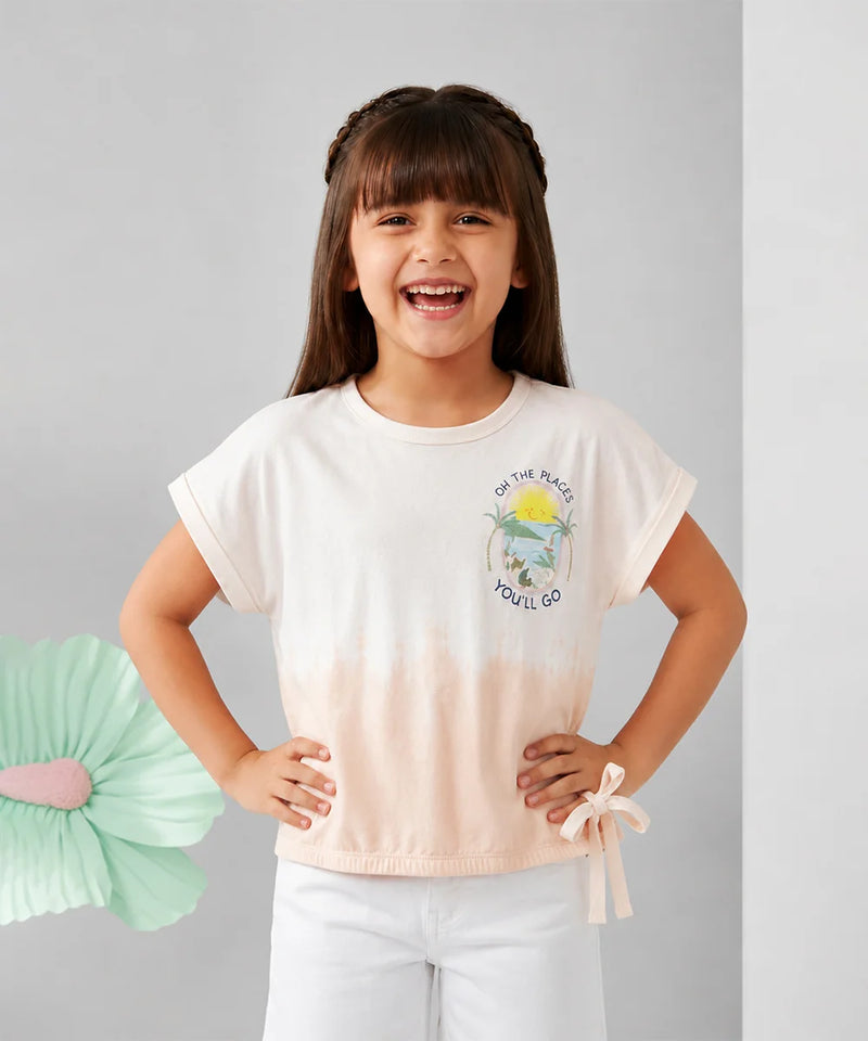 A young girl with dark hair in braids smiling, wearing a short-sleeve tie-dye t-shirt with a small graphic on the chest and white pants, standing against a light gray background.