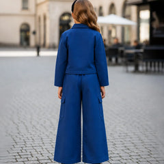 A woman viewed from behind wearing a bright royal blue matching set consisting of a cropped long-sleeve jacket and wide-leg trousers, standing on a cobblestone street in an outdoor urban setting.
