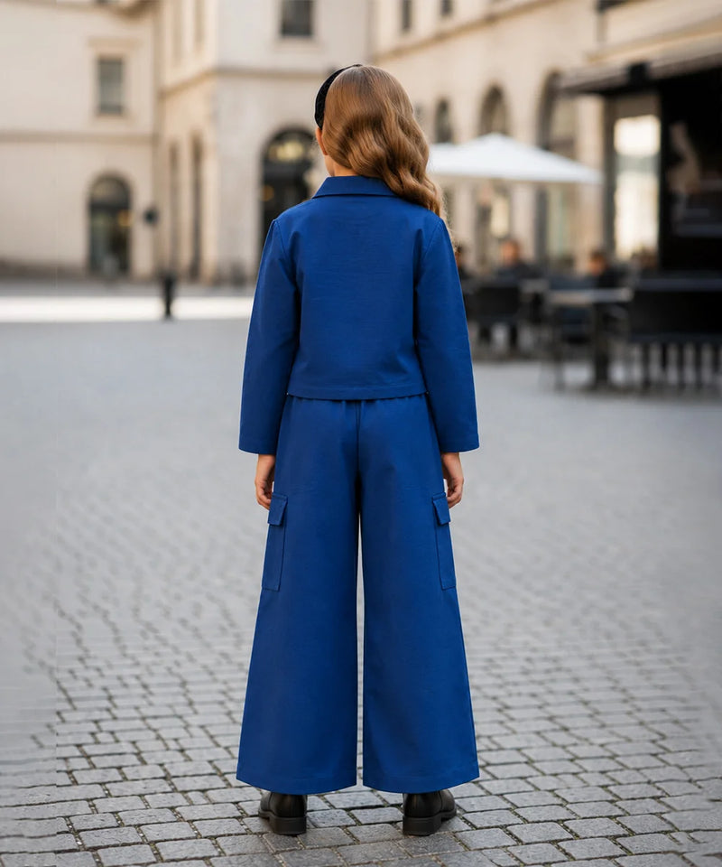 A woman viewed from behind wearing a bright royal blue matching set consisting of a cropped long-sleeve jacket and wide-leg trousers, standing on a cobblestone street in an outdoor urban setting.