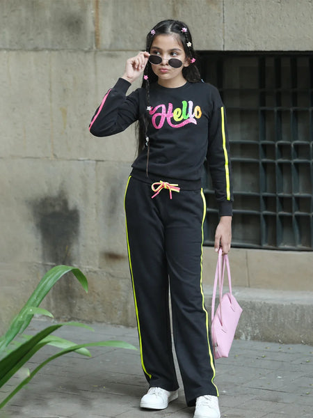 A woman modeling a black tracksuit with neon yellow and pink details, featuring a 'Hello' graphic on the chest, paired with white sneakers and a pink handbag.