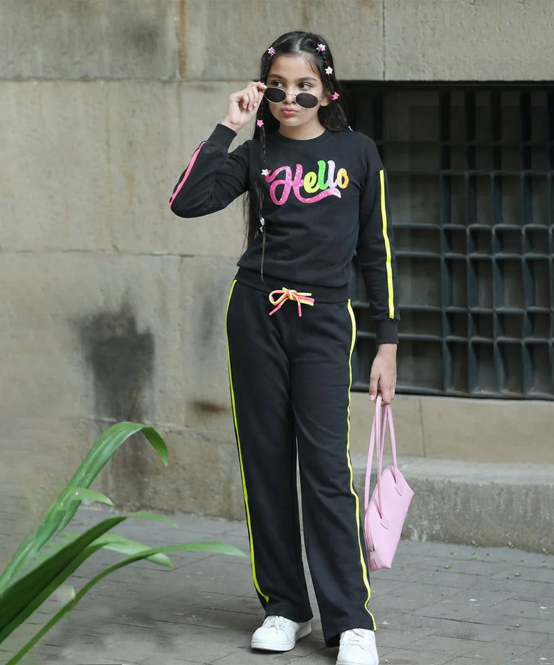 A woman modeling a black tracksuit with neon yellow and pink details, featuring a 'Hello' graphic on the chest, paired with white sneakers and a pink handbag.