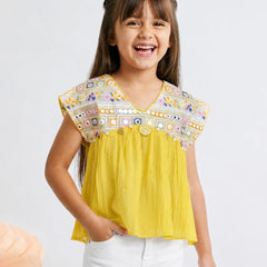 A young girl smiling and wearing a bright yellow sleeveless top with a heavily embroidered multicolor yoke and white pants, photographed against a light gray studio background.