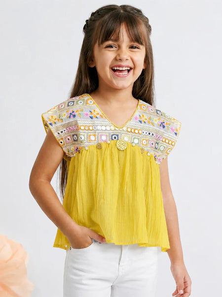 A young girl smiling and wearing a bright yellow sleeveless top with a heavily embroidered multicolor yoke and white pants, photographed against a light gray studio background.