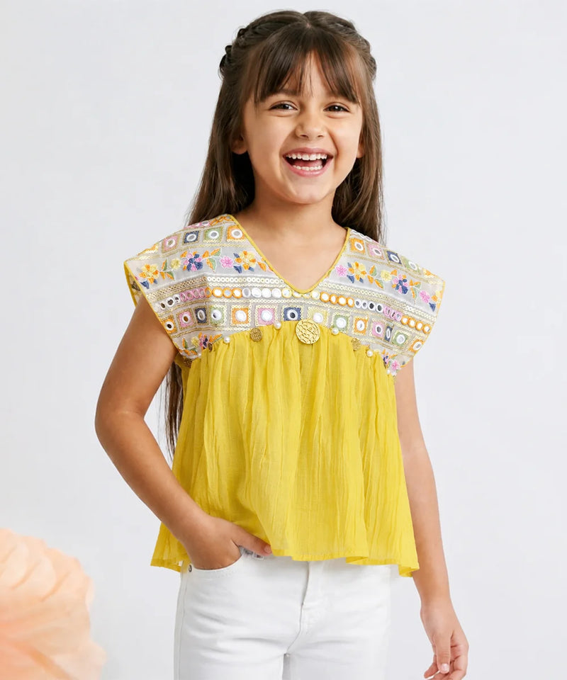 A young girl smiling and wearing a bright yellow sleeveless top with a heavily embroidered multicolor yoke and white pants, photographed against a light gray studio background.