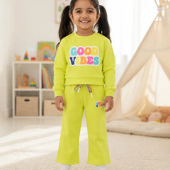 A young girl stands in a brightly lit children's room wearing a matching neon yellow-green sweatshirt and sweatpants set. The sweatshirt features the words 'GOOD VIBES' in colorful bubble letters, and the pants have a small matching logo patch on the thigh.