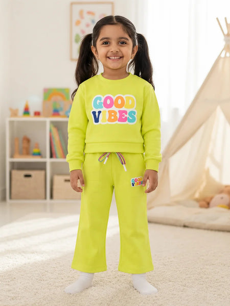A young girl stands in a brightly lit children's room wearing a matching neon yellow-green sweatshirt and sweatpants set. The sweatshirt features the words 'GOOD VIBES' in colorful bubble letters, and the pants have a small matching logo patch on the thigh.