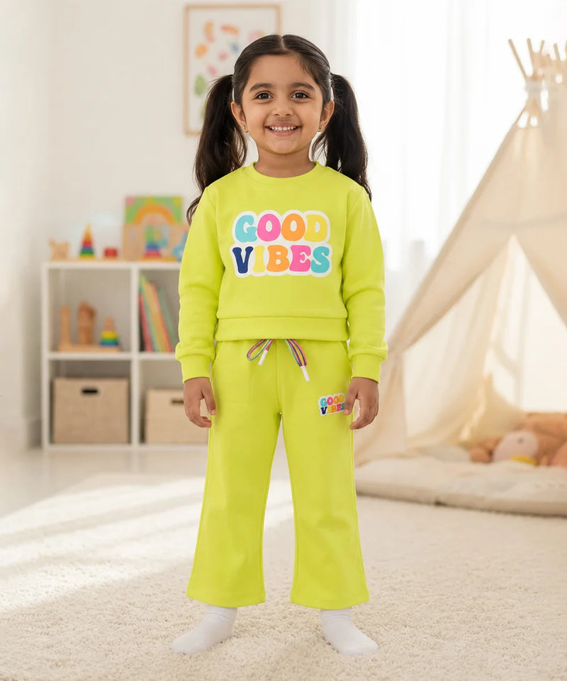 A young girl stands in a brightly lit children's room wearing a matching neon yellow-green sweatshirt and sweatpants set. The sweatshirt features the words 'GOOD VIBES' in colorful bubble letters, and the pants have a small matching logo patch on the thigh.
