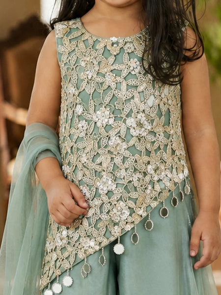 A child wearing a sleeveless mint green and beige embroidered traditional outfit with intricate lace embroidery, white floral appliques, and dangling tassel details, paired with a matching sheer dupatta.
