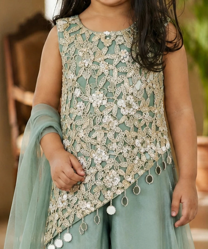 A child wearing a sleeveless mint green and beige embroidered traditional outfit with intricate lace embroidery, white floral appliques, and dangling tassel details, paired with a matching sheer dupatta.