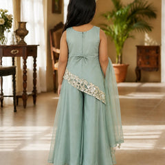A young girl viewed from behind wearing a sage green sleeveless gown with a matching sheer dupatta and a decorative embroidered sash, standing in a warmly lit indoor room.