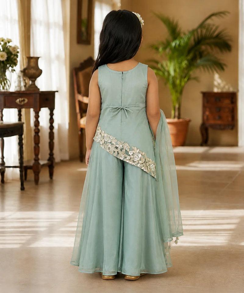 A young girl viewed from behind wearing a sage green sleeveless gown with a matching sheer dupatta and a decorative embroidered sash, standing in a warmly lit indoor room.