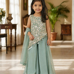 A young girl stands in a warmly lit indoor room wearing a mint green traditional outfit with a heavily embroidered light beige and silver sleeveless top, matching wide-leg pants, and a sheer dupatta.