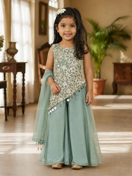 A young girl stands in a warmly lit indoor room wearing a mint green traditional outfit with a heavily embroidered light beige and silver sleeveless top, matching wide-leg pants, and a sheer dupatta.