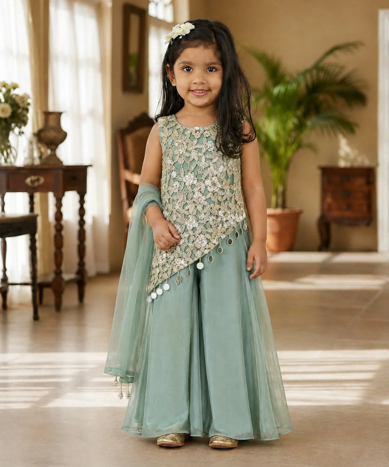 A young girl stands in a warmly lit indoor room wearing a mint green traditional outfit with a heavily embroidered light beige and silver sleeveless top, matching wide-leg pants, and a sheer dupatta.