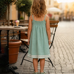 A young girl with blonde hair is seen from behind, wearing a light mint green sleeveless dress with a white lace hem, standing on a cobblestone street in an outdoor cafe setting.