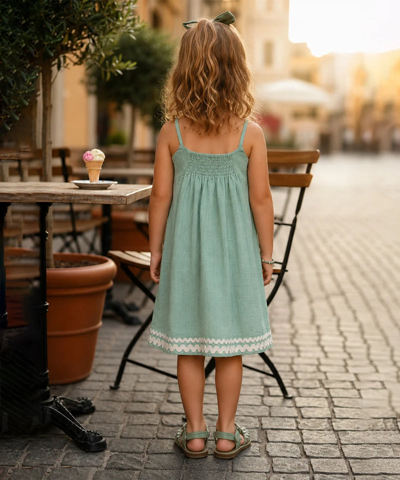 A young girl with blonde hair is seen from behind, wearing a light mint green sleeveless dress with a white lace hem, standing on a cobblestone street in an outdoor cafe setting.