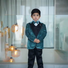 A boy wearing a green tuxedo with a white shirt, black waistcoat, and black pants. He has a matching bow tie, broach, and pocket square.