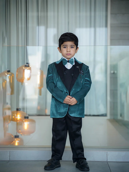 A boy wearing a green tuxedo with a white shirt, black waistcoat, and black pants. He has a matching bow tie, broach, and pocket square.