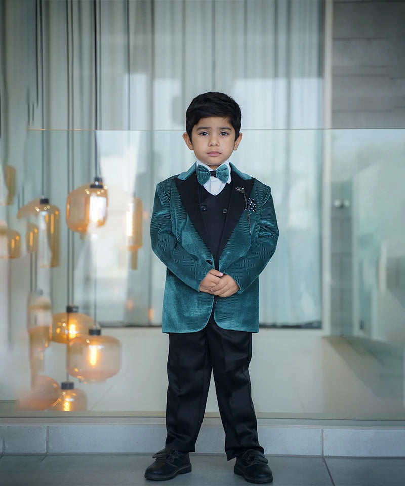 A boy wearing a green tuxedo with a white shirt, black waistcoat, and black pants. He has a matching bow tie, broach, and pocket square.