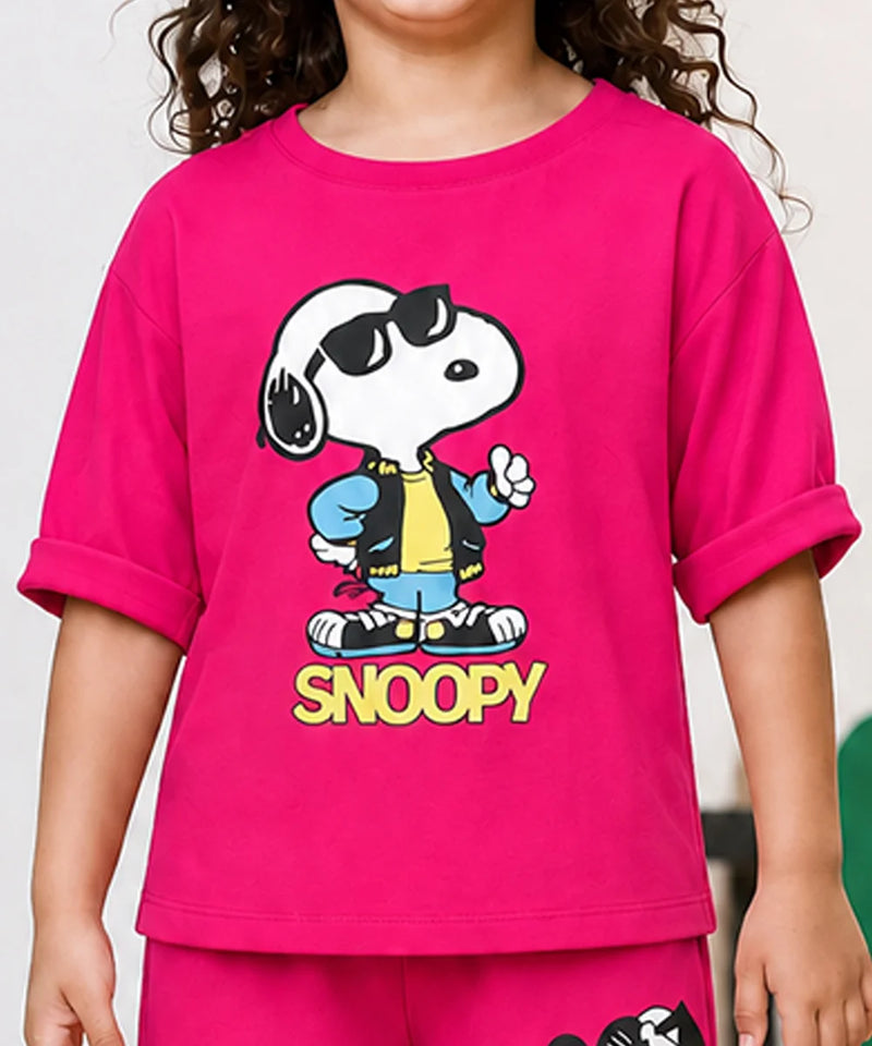 A child wearing a bright pink short-sleeve T-shirt with a large Snoopy graphic on the front. The graphic shows Snoopy wearing black sunglasses, a blue jacket, a yellow shirt, and blue jeans, with the word 'SNOOPY' printed below in yellow.