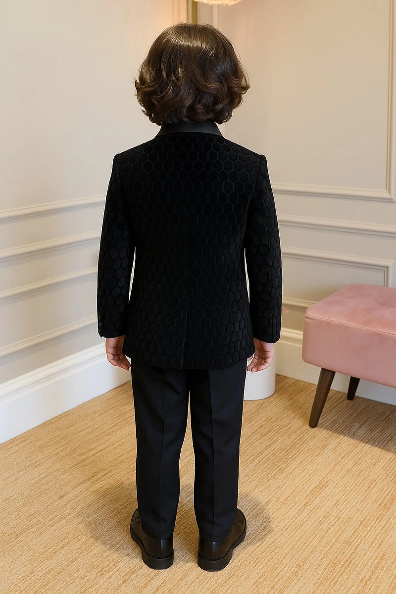 Person wearing a black textured jacket and pants standing in a room with a pink stool and white walls.