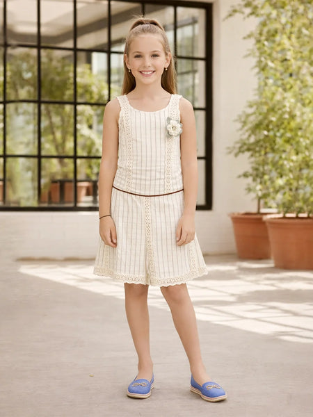 Young girl in a white dress standing in a sunlit room with large windows and plants.