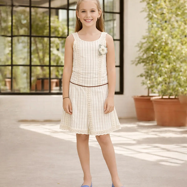Young girl in a white dress standing in a sunlit room with large windows and plants.