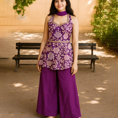 Woman wearing a purple floral top and pants standing outdoors with a bench and greenery in the background.