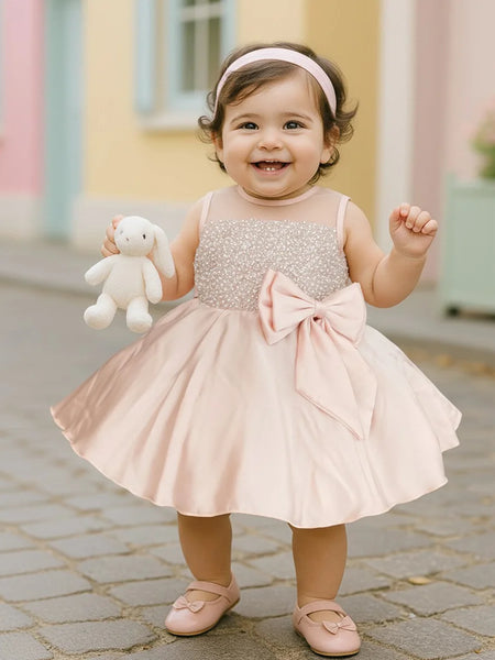 Child in a sparkly dress with a large bow holding a stuffed toy outdoors.