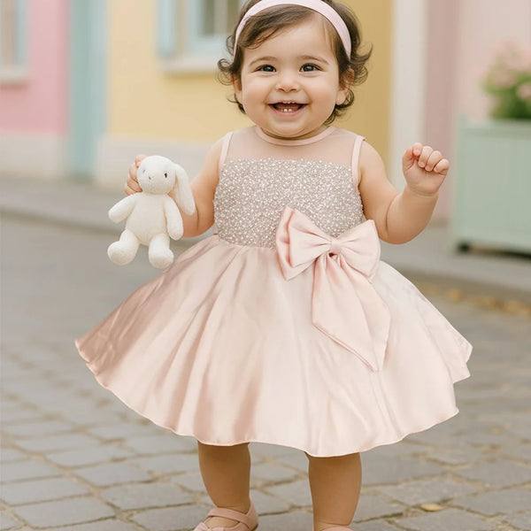 Child in a sparkly dress with a large bow holding a stuffed toy outdoors.