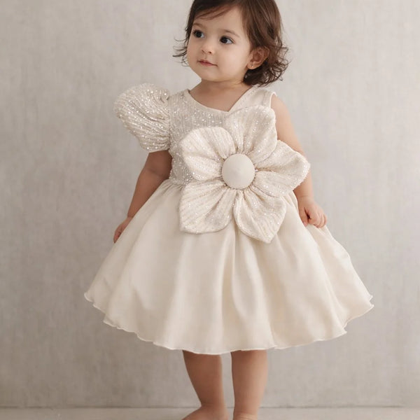 Young child wearing a white dress with a large flower detail against a neutral background