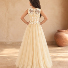 A young girl viewed from behind wearing a formal cream tulle ball gown with an intricate lace and applique bodice, standing in a warm, rustic room with a large clay pot in the background.