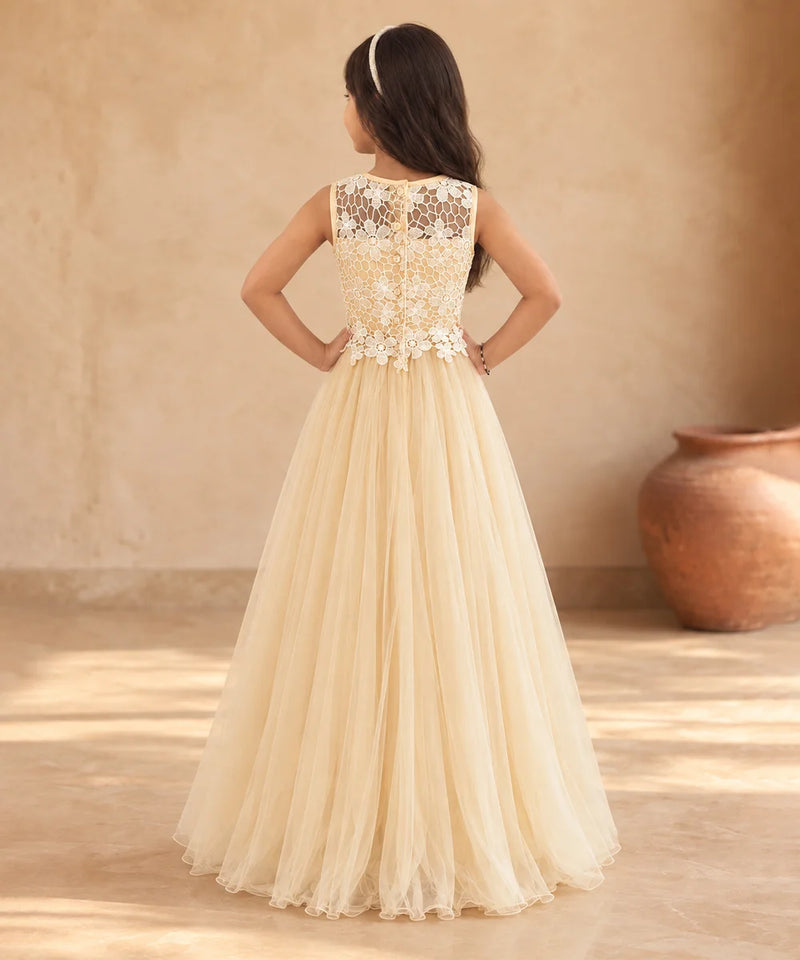 A young girl viewed from behind wearing a formal cream tulle ball gown with an intricate lace and applique bodice, standing in a warm, rustic room with a large clay pot in the background.