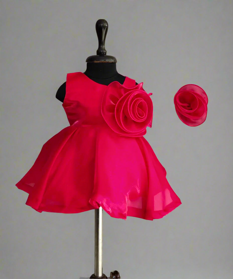 A red colored kids' party frock displayed on a mannequin, featuring a back zip closure, floral detailing on the waist, and paired with a matching hair accessory.