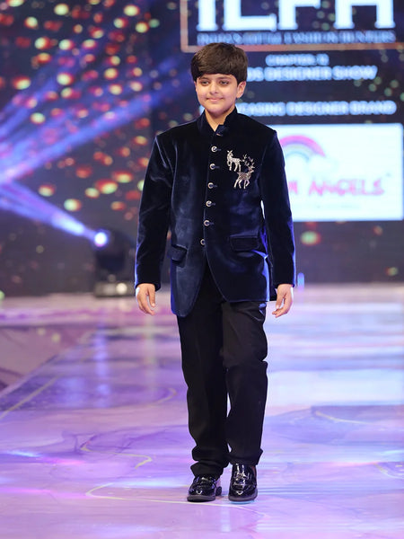 Young boy in a formal outfit walking on a stage with colorful lights and branding in the background.