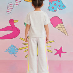 A child viewed from behind wearing a light heather gray short-sleeve t-shirt and matching wide-leg pants with yellow and pink side stripes, standing against a pastel pink wall decorated with cartoon beach and summer motifs.