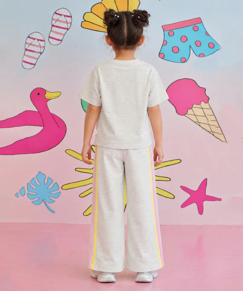 A child viewed from behind wearing a light heather gray short-sleeve t-shirt and matching wide-leg pants with yellow and pink side stripes, standing against a pastel pink wall decorated with cartoon beach and summer motifs.