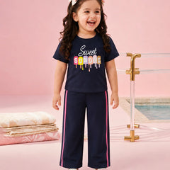 A young girl with curly brown hair wearing a navy blue two-piece pajama set with a colorful 'Sweet Sorority' graphic on the t-shirt and pink side stripes on the pants, standing in a pink studio setting with a pool background.