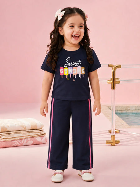 A young girl with curly brown hair wearing a navy blue two-piece pajama set with a colorful 'Sweet Sorority' graphic on the t-shirt and pink side stripes on the pants, standing in a pink studio setting with a pool background.