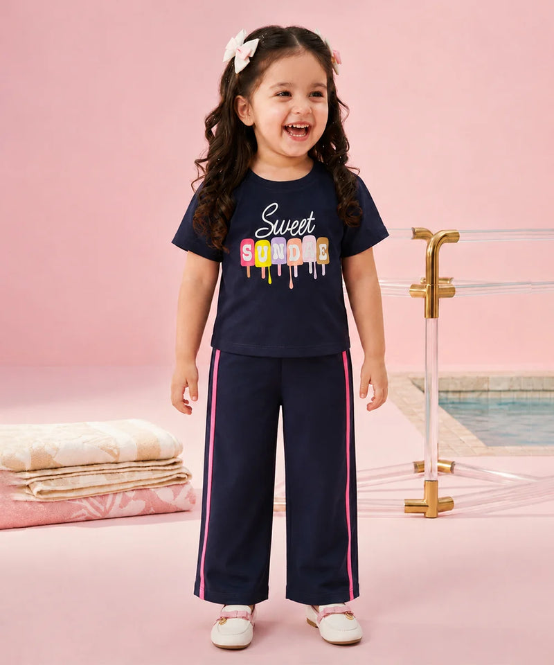 A young girl with curly brown hair wearing a navy blue two-piece pajama set with a colorful 'Sweet Sorority' graphic on the t-shirt and pink side stripes on the pants, standing in a pink studio setting with a pool background.
