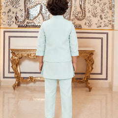 A child viewed from behind wearing a light mint green two-piece traditional outfit with a short kurta and trousers, standing in front of an ornate gold console table and a decorated wall.