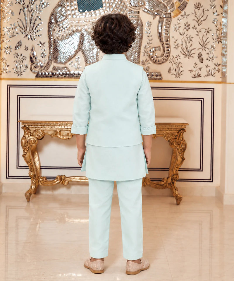 A child viewed from behind wearing a light mint green two-piece traditional outfit with a short kurta and trousers, standing in front of an ornate gold console table and a decorated wall.