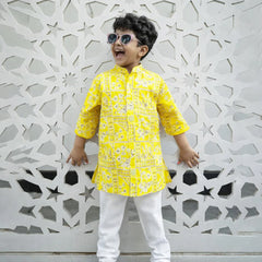 Child wearing a yellow kurta with white pants standing against an ornate white wall.