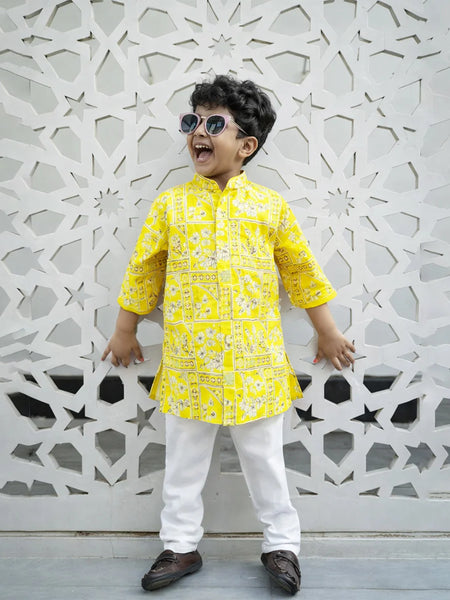 Child wearing a yellow kurta with white pants standing against an ornate white wall.