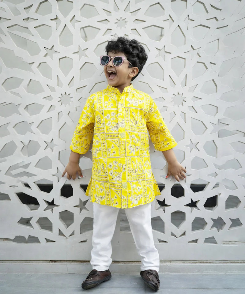 Child wearing a yellow kurta with white pants standing against an ornate white wall.