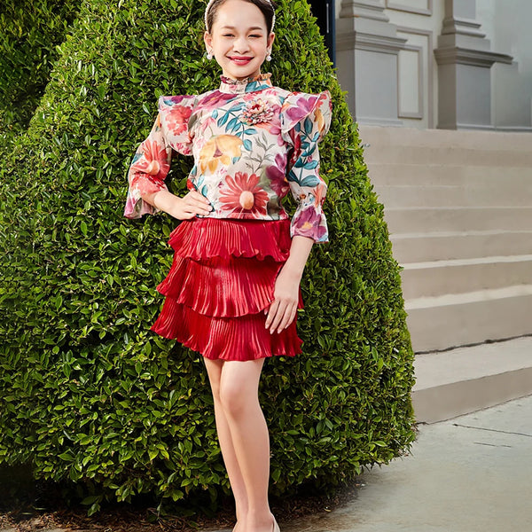 Woman in a floral top and red skirt standing next to a green bush